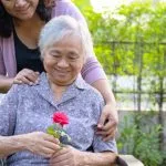 Older woman in chair smiling at a rose while younger woman embraces her from behind.